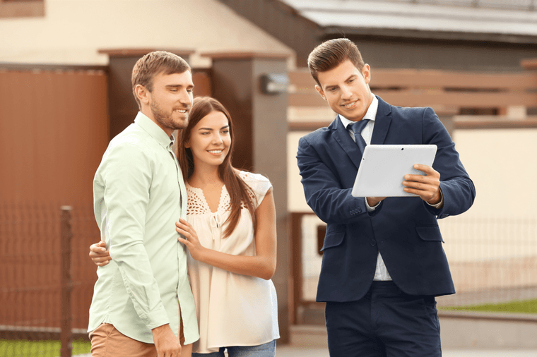 A real estate agent in a blue suit shows a tablet to a smiling couple in front of a house.