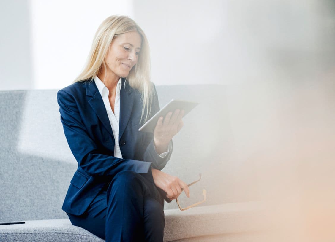 Woman in a navy suit sits on a gray sofa, smiling while looking at a tablet. She holds glasses in her other hand.