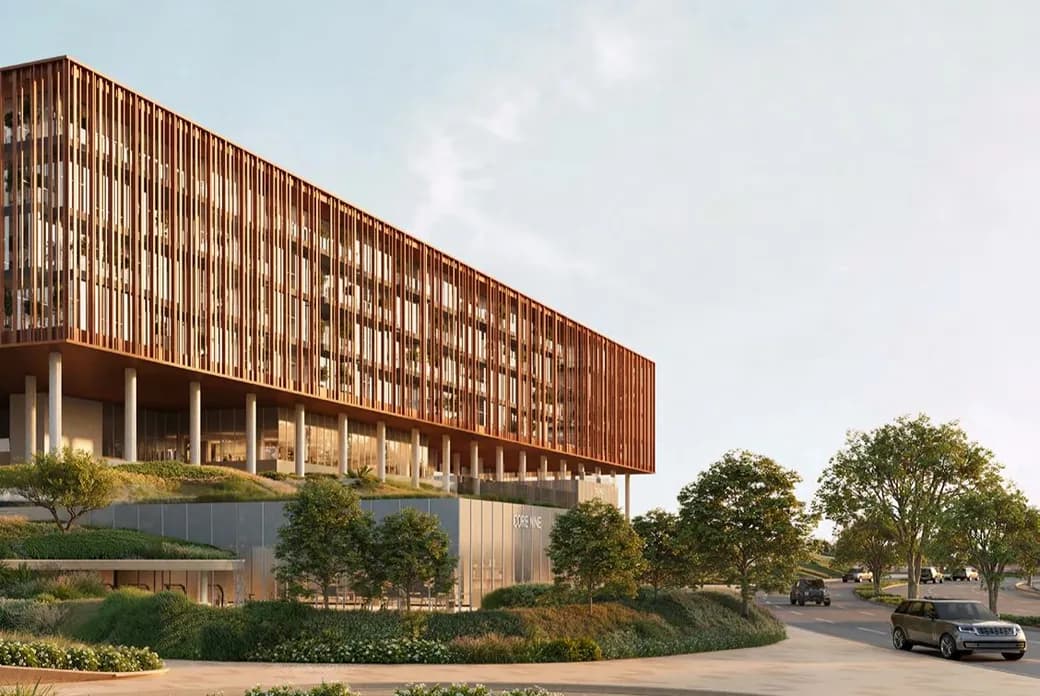 Contemporary glass-fronted building with a vertical wooden slat façade, elevated on columns, surrounded by trees and a curved road with cars.