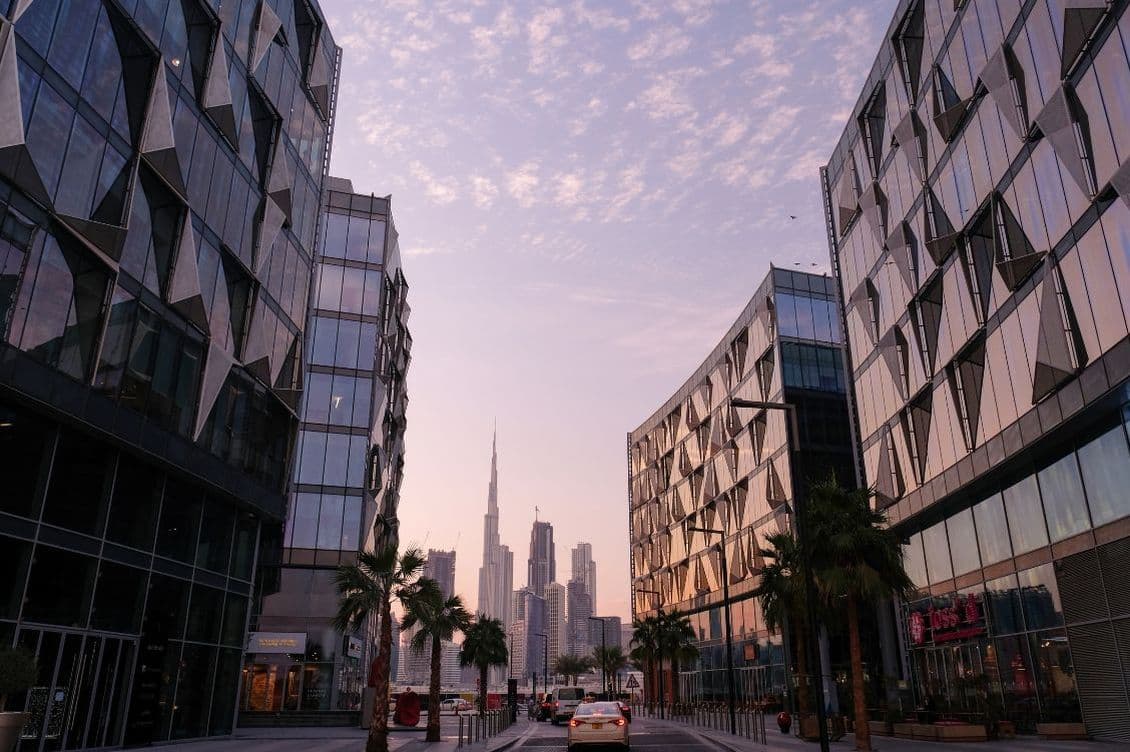 Modern cityscape with tall glass buildings on both sides of a street, leading to a distant skyline under a pastel sky at dusk.
