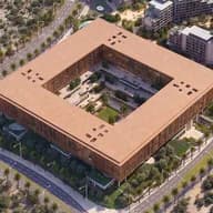 Aerial view of a large square building with an open central courtyard, brown facade, surrounded by palm-lined streets and modern apartments.
