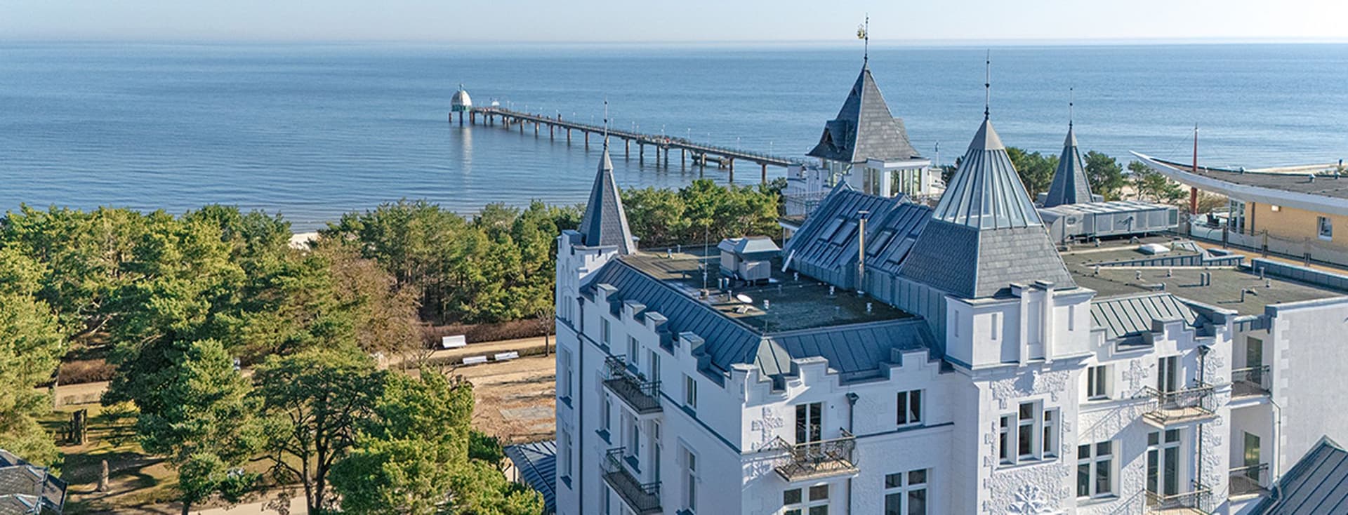Ein weißes Hotel mit Türmchen am Meer, mit Blick auf eine von Bäumen gesäumte Küste und einen langen Steg, der sich in das ruhige Meer erstreckt
