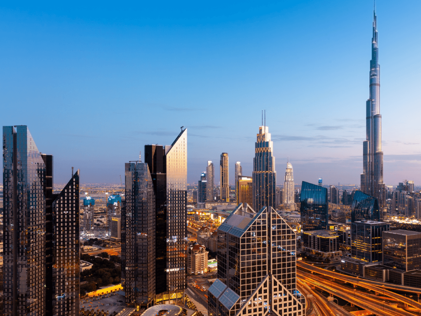 Skyline of Dubai at dusk, featuring illuminated skyscrapers, including a prominent tall building, with a network of roads below.