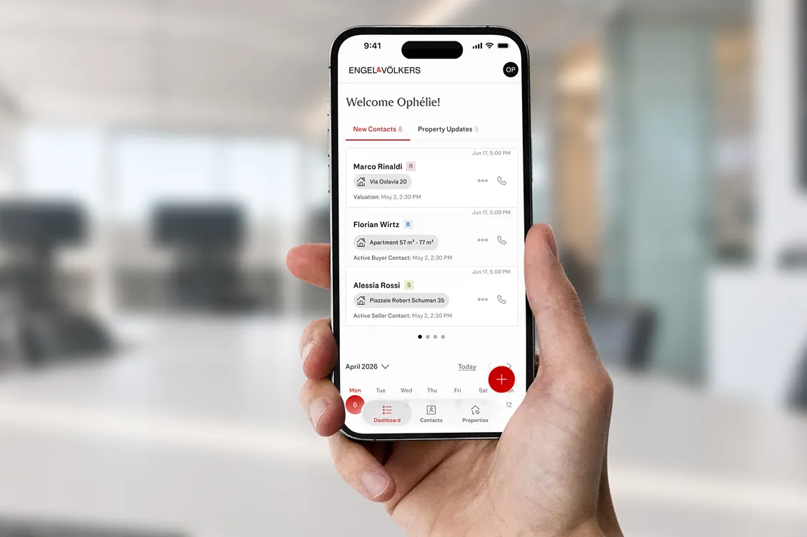 Hand holding a smartphone displaying a Engel & Völkers Advance app dashboard with new contacts and property updates; blurred office background. AI-generated image