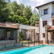 Luxury multi-level villa with tiled roofs, stone pillars, outdoor dining patio, swimming pool with glass railing, and surrounding lush trees.