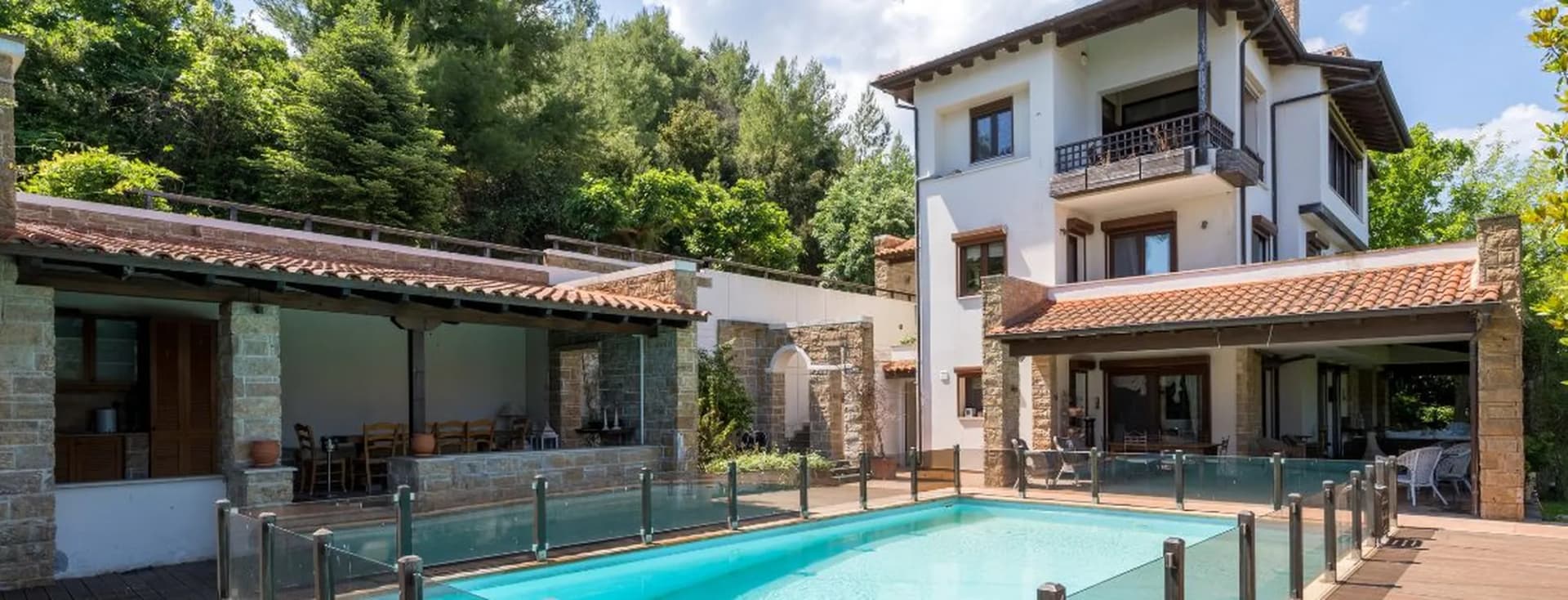 Luxury multi-level villa with tiled roofs, stone pillars, outdoor dining patio, swimming pool with glass railing, and surrounding lush trees.