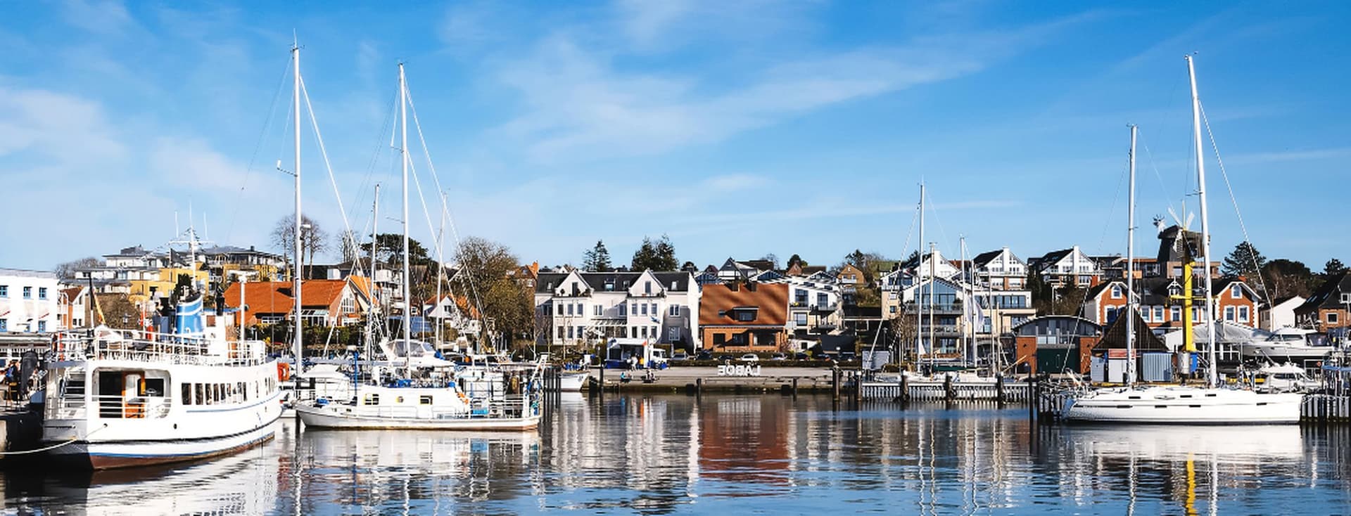 Der sonnige Hafen von Laboe mit Segelbooten und Yachten, die vor bunten Häusern am Wasser vor Anker liegen, blauer Himmel und Spiegelungen auf dem ruhigen Wasser