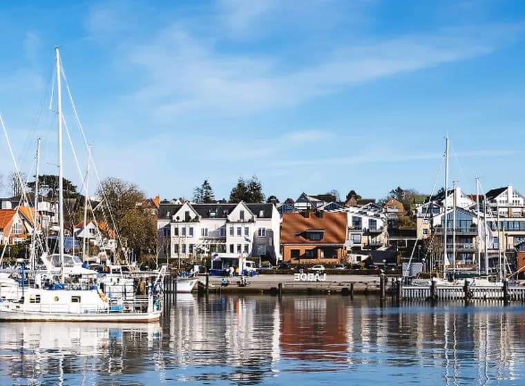Der sonnige Hafen von Laboe mit Segelbooten und Yachten, die vor bunten Häusern am Wasser vor Anker liegen, blauer Himmel und Spiegelungen auf dem ruhigen Wasser