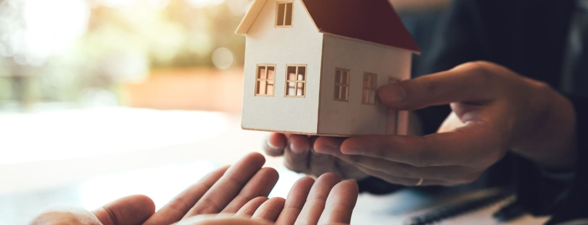 Hands handling a small model house to open hands, symbolizing a home transfer, sale, or real estate agreement.