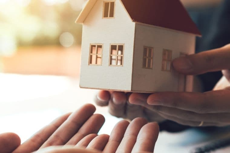Hands handing a small model house to open hands, symbolizing a home transfer, sale, or real estate agreement.