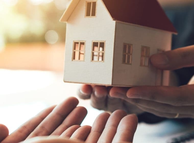 Hands handing a small model house to open hands, symbolizing a home transfer, sale, or real estate agreement.