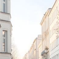 Sunlit street of pale buildings with a black-railed balcony on the left and a blooming cherry tree on the right.