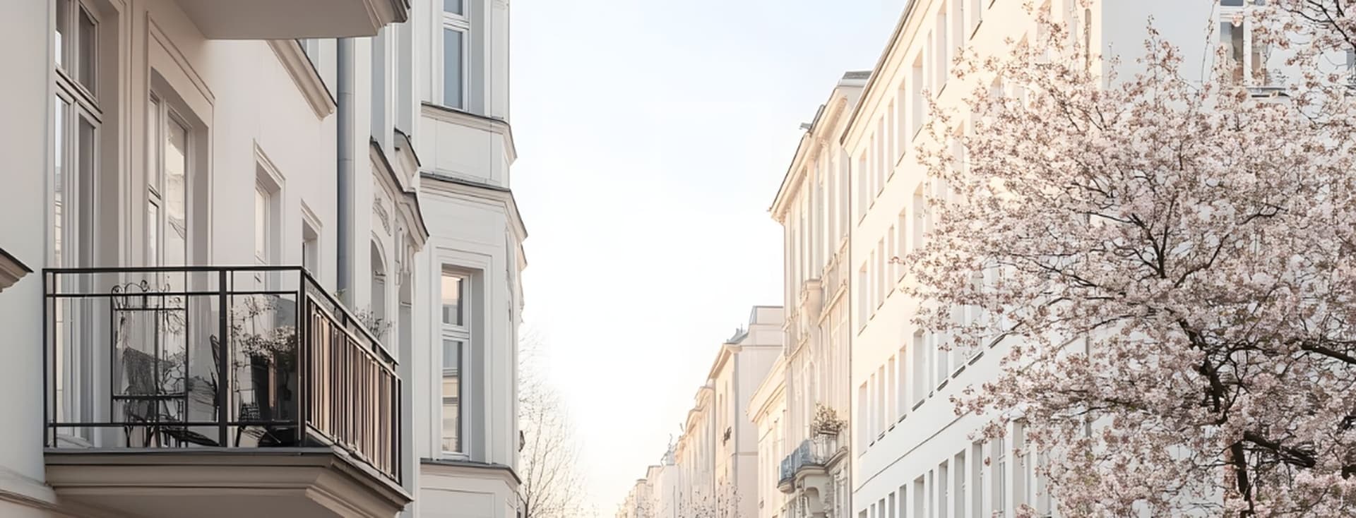 Sunlit street of pale buildings with a black-railed balcony on the left and a blooming cherry tree on the right.