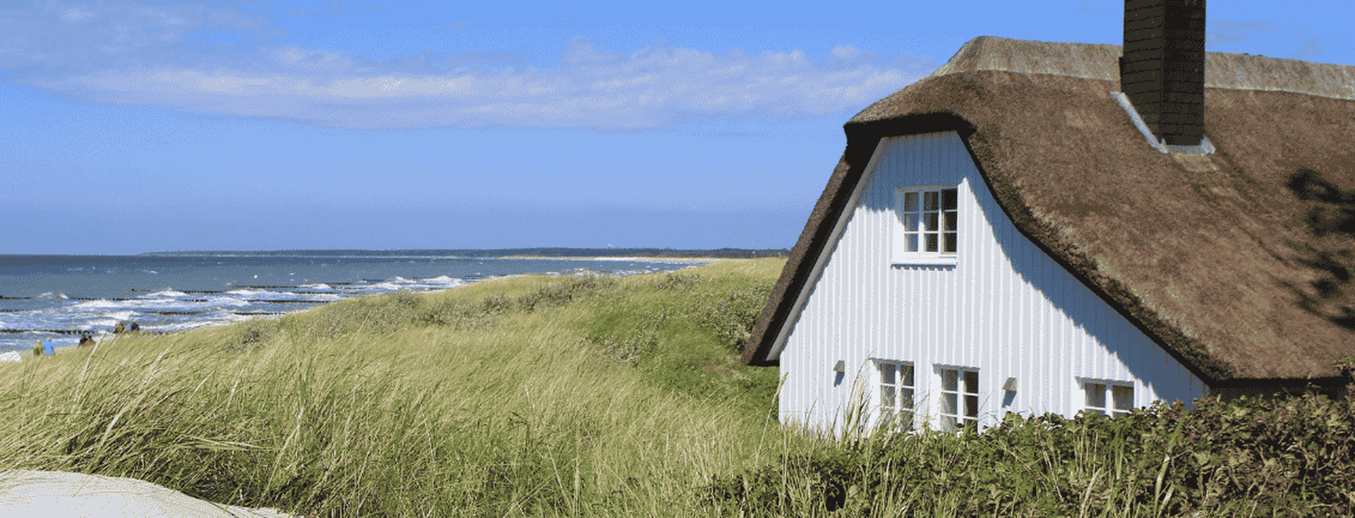 Weißes Häuschen mit Strohdach inmitten von Küstendünen mit hohem Gras, mit Blick auf das Meer unter blauem Himmel