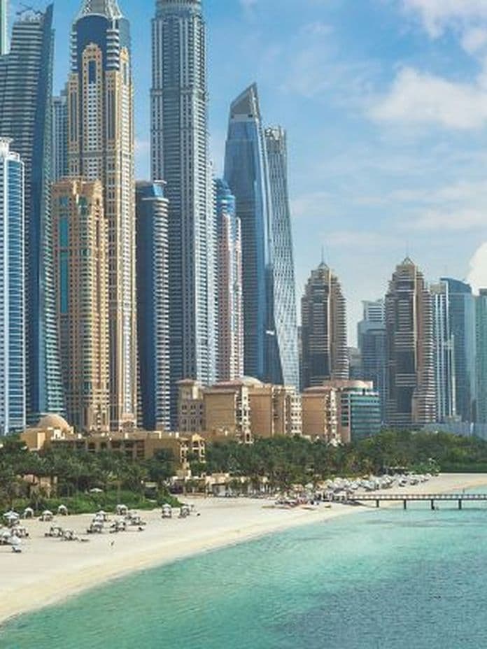 Skyline of tall, modern skyscrapers by a sandy beach and turquoise sea under a partly cloudy sky, with palm trees and beach umbrellas in the foreground.