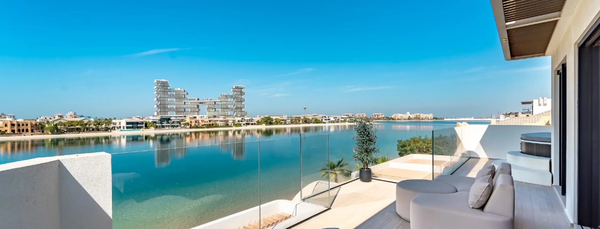 Modern balcony view with sleek glass railing overlooking a calm waterfront and distinctive, futuristic buildings under a clear blue sky.
