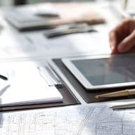 Close-up of two professionals' hands reviewing architectural plans and charts with tablet, clipboard, pen, and eyeglasses on desk.