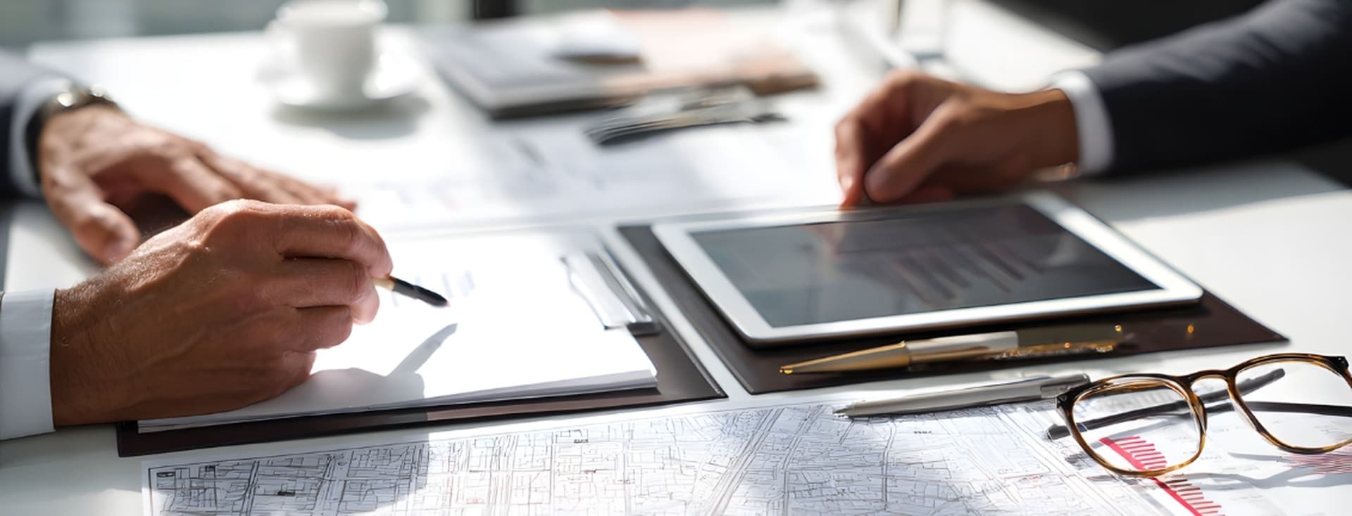 Close-up of two professionals' hands reviewing architectural plans and charts with tablet, clipboard, pen, and eyeglasses on desk.