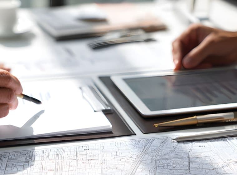 Close-up of two professionals' hands reviewing architectural plans and charts with tablet, clipboard, pen, and eyeglasses on desk.