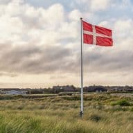 Eine dänische Flagge weht über grasbewachsenen Küstendünen mit strohgedeckten Häuschen unter einem bewölkten Himmel