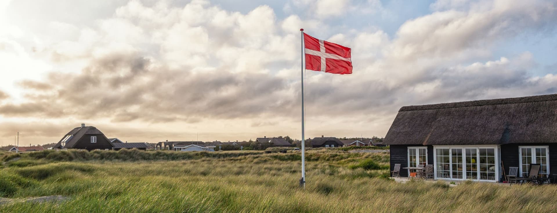 Eine dänische Flagge weht über grasbewachsenen Küstendünen mit strohgedeckten Häuschen unter einem bewölkten Himmel