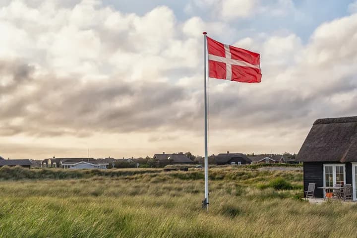 Eine dänische Flagge weht über grasbewachsenen Küstendünen mit strohgedeckten Häuschen unter einem bewölkten Himmel