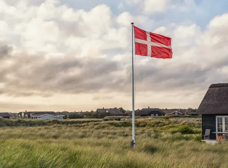 Eine dänische Flagge weht über grasbewachsenen Küstendünen mit strohgedeckten Häuschen unter einem bewölkten Himmel