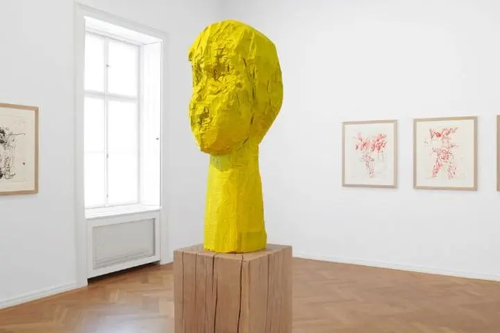 Bright gallery space with yellow wooden sculpture on a pedestal, parquet flooring and three drawings on white walls