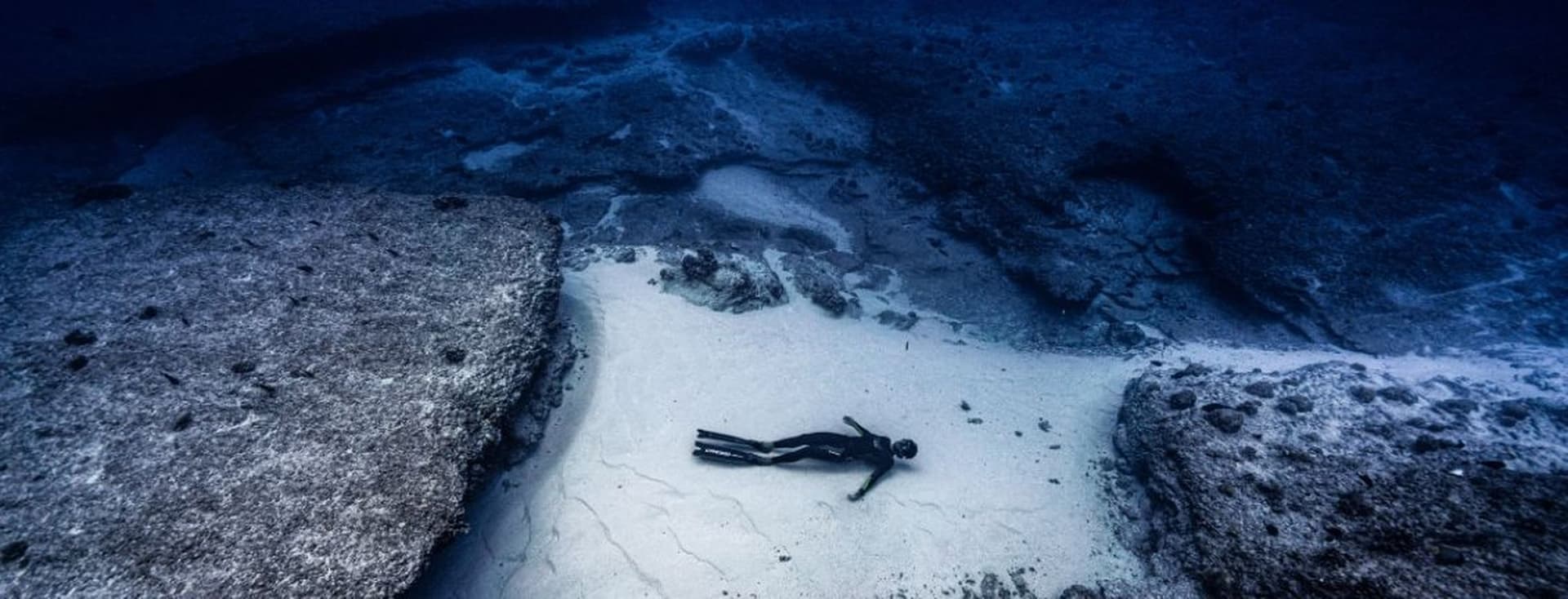 Ein Freitaucher im Neoprenanzug liegt auf dem sandigen Meeresgrund zwischen Felsformationen im tiefblauen Wasser.