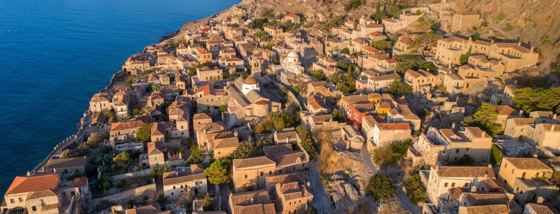 Aerial view of the medieval stone castle town of Monemvasia with brown-tiled roofs overlooking the deep blue sea.