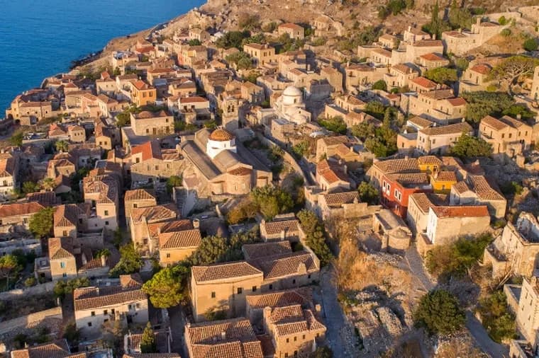 Aerial view of the medieval stone castle town of Monemvasia with brown-tiled roofs overlooking the deep blue sea.