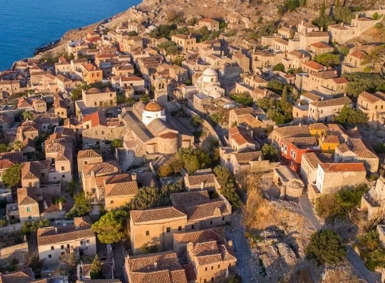 Aerial view of the medieval stone castle town of Monemvasia with brown-tiled roofs overlooking the deep blue sea.
