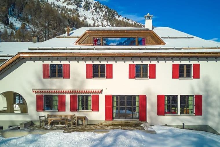 Large white Alpine house with red shutters and a terrace, surrounded by snow-covered fir trees and mountains under a bright blue sky.