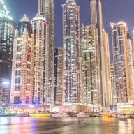 Night view of Dubai Marina with tall skyscrapers reflecting on the water, boats docked along a lively waterfront.