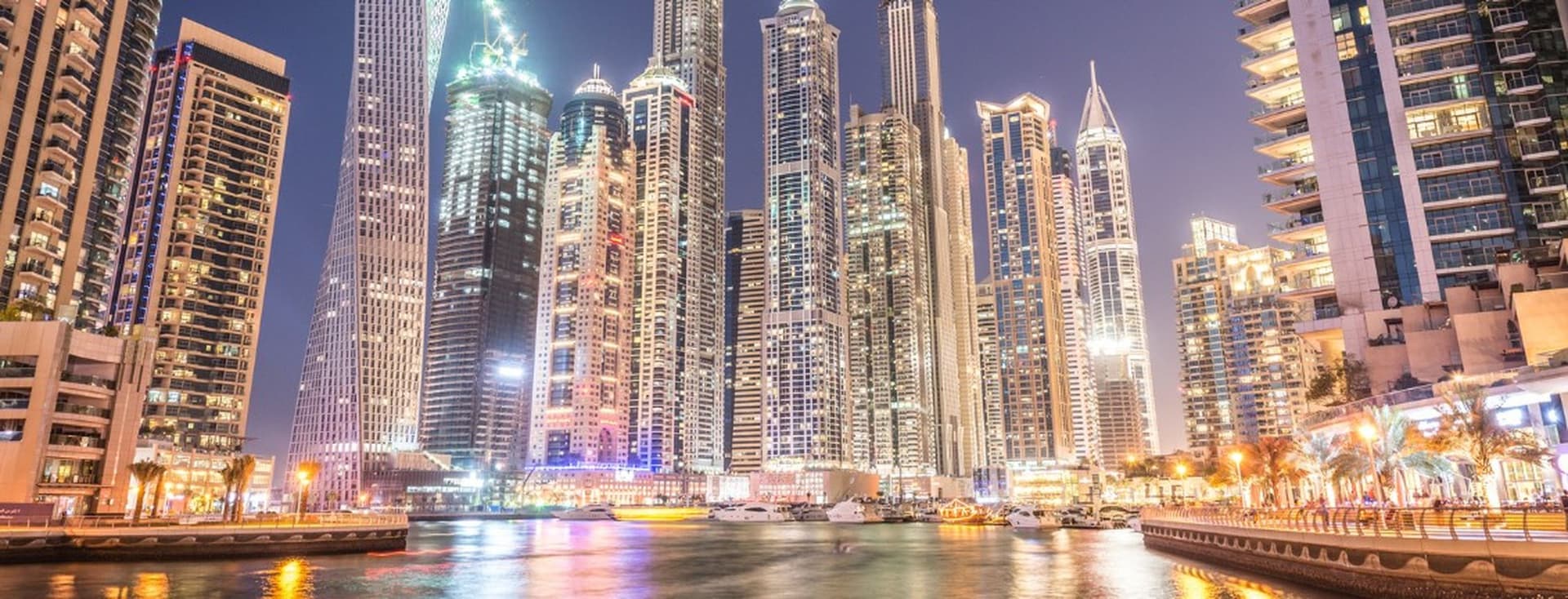 Night view of Dubai Marina with tall skyscrapers reflecting on the water, boats docked along a lively waterfront.