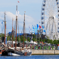 Historische Segelschiffe, die bei einem belebten Festival am Wasser vor Anker liegen, mit bunten Fahnen, Menschenmengen, weißen Zelten und einem großen Riesenrad