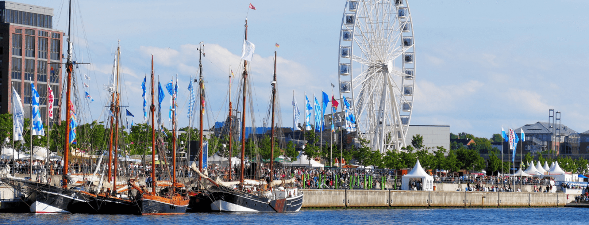 Historische Segelschiffe, die bei einem belebten Festival am Wasser vor Anker liegen, mit bunten Fahnen, Menschenmengen, weißen Zelten und einem großen Riesenrad