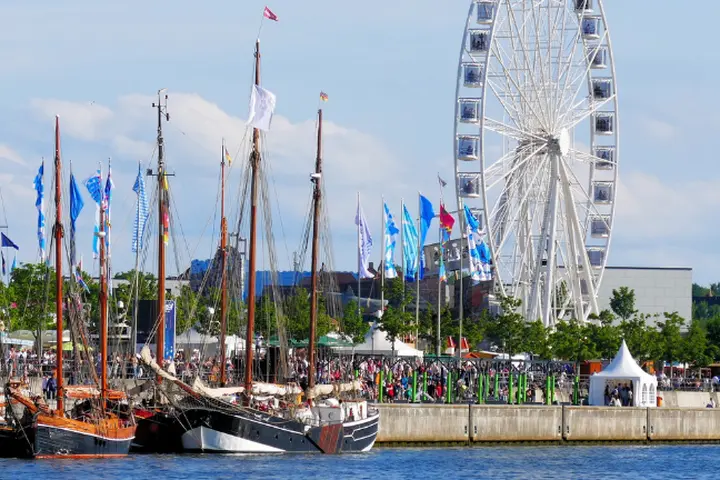 Historische Segelschiffe, die bei einem belebten Festival am Wasser vor Anker liegen, mit bunten Fahnen, Menschenmengen, weißen Zelten und einem großen Riesenrad