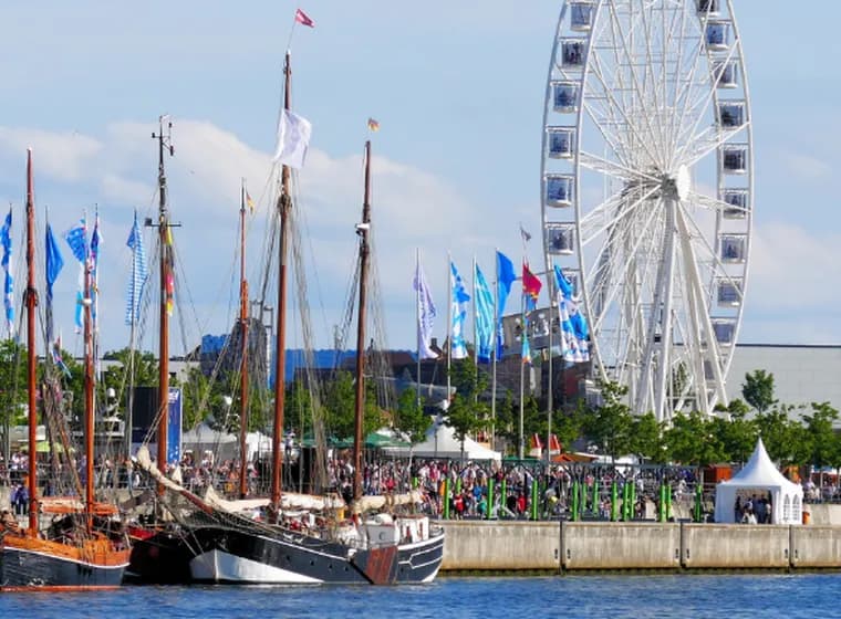 Historische Segelschiffe, die bei einem belebten Festival am Wasser vor Anker liegen, mit bunten Fahnen, Menschenmengen, weißen Zelten und einem großen Riesenrad