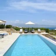 Modern villa terrace with long lap pool, daybeds, umbrellas, and scenic ocean backdrop.