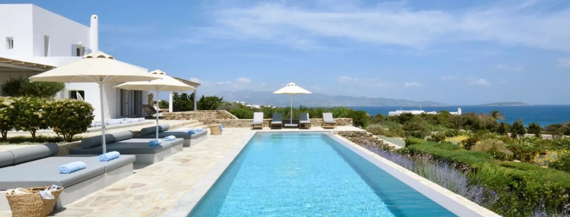 Modern villa terrace with long lap pool, daybeds, umbrellas, and scenic ocean backdrop.
