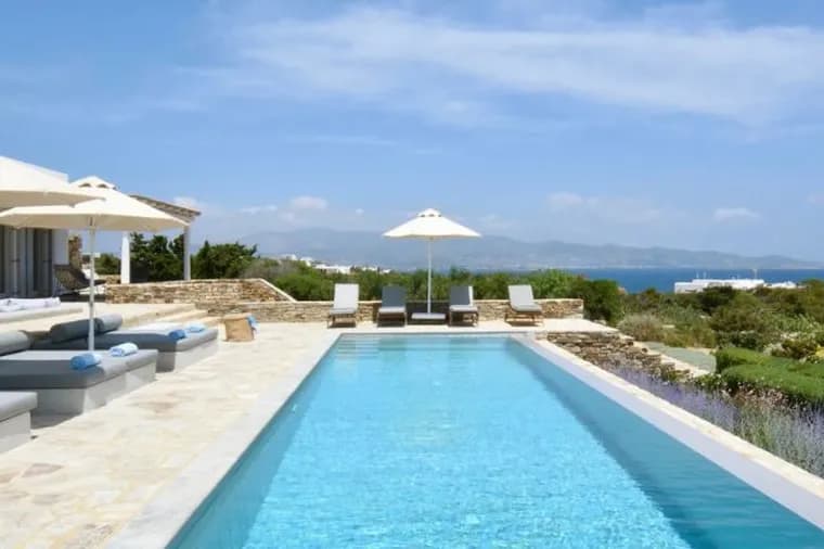 Modern villa terrace with long lap pool, daybeds, umbrellas, and scenic ocean backdrop.