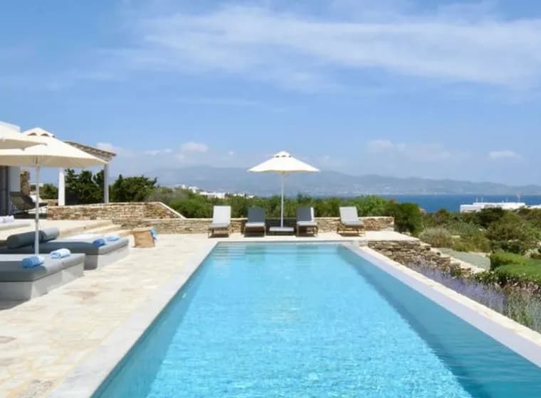 Modern villa terrace with long lap pool, daybeds, umbrellas, and scenic ocean backdrop.