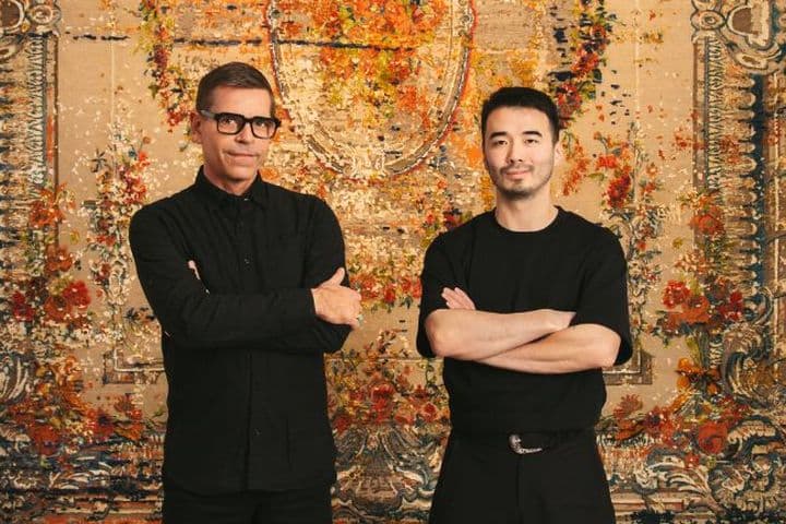 Zwei Männer stehen mit verschränkten Armen vor einem kunstvollen, farbenfrohen Wandteppich. Beide tragen dunkle Kleidung und Brillen und blicken selbstbewusst in die Kamera.