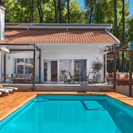 Luxury villa with a private swimming pool surrounded by pine trees. The property features a white exterior, terracotta roof tiles, a spacious wooden deck, sun loungers with umbrellas, and outdoor lounge areas, creating a tranquil resort-style setting.