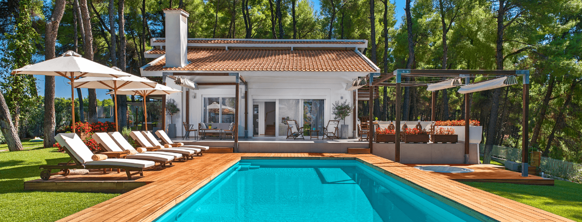 Luxury villa with a private swimming pool surrounded by pine trees. The property features a white exterior, terracotta roof tiles, a spacious wooden deck, sun loungers with umbrellas, and outdoor lounge areas, creating a tranquil resort-style setting.