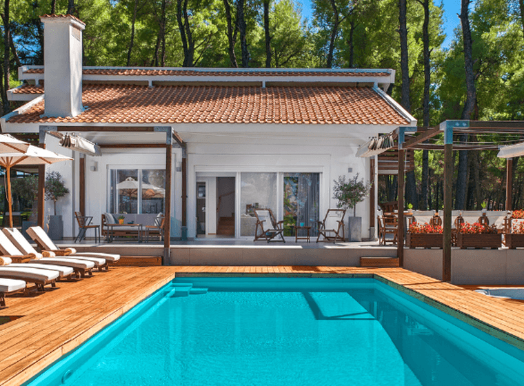 Luxury villa with a private swimming pool surrounded by pine trees. The property features a white exterior, terracotta roof tiles, a spacious wooden deck, sun loungers with umbrellas, and outdoor lounge areas, creating a tranquil resort-style setting.