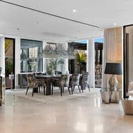 Modern open-concept living area with a kitchen, bar seating, dining table, and sofa. Large windows reveal a pool and palm trees outside.