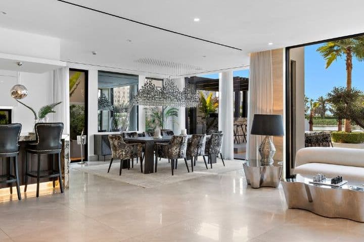 Modern open-concept living area with a kitchen, bar seating, dining table, and sofa. Large windows reveal a pool and palm trees outside.