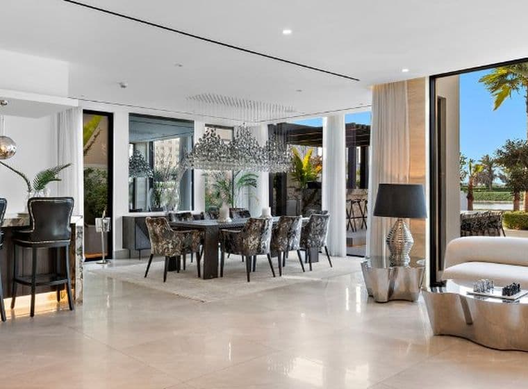 Modern open-concept living area with a kitchen, bar seating, dining table, and sofa. Large windows reveal a pool and palm trees outside.